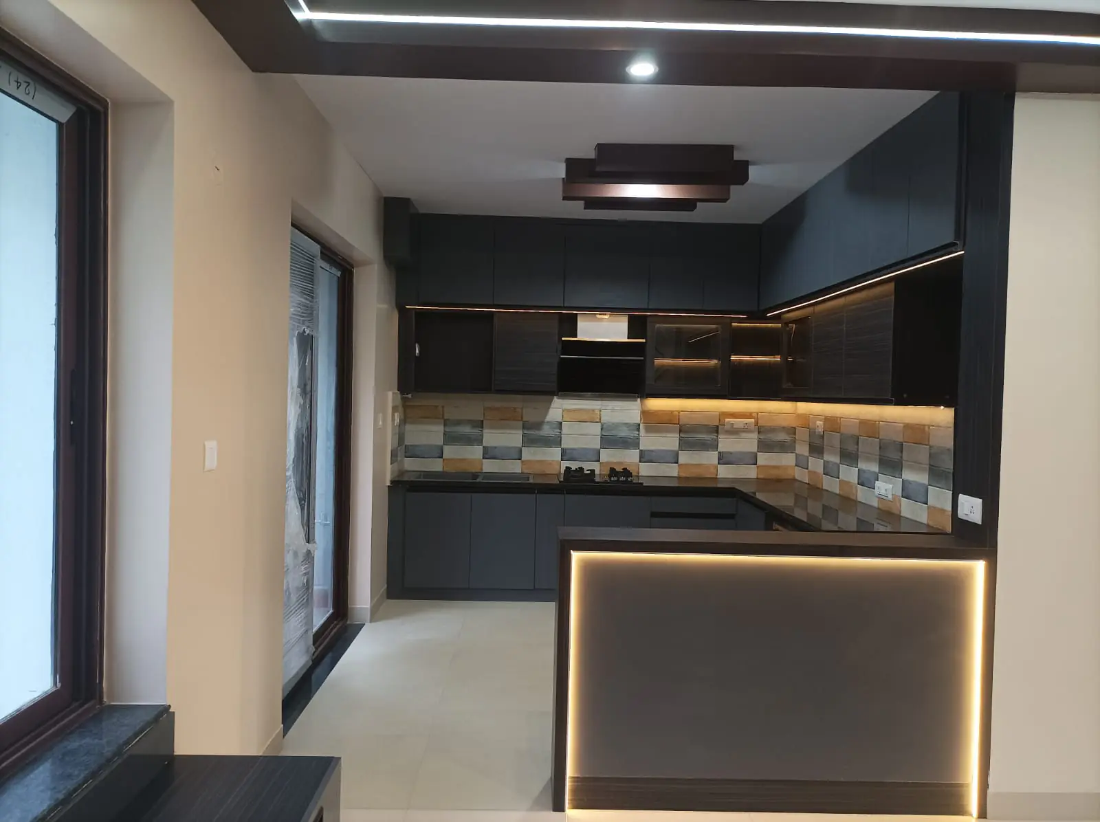 A finished modular kitchen in dark shutters, with a tiled backsplash, under-cabinet lighting and a lit breakfast counter