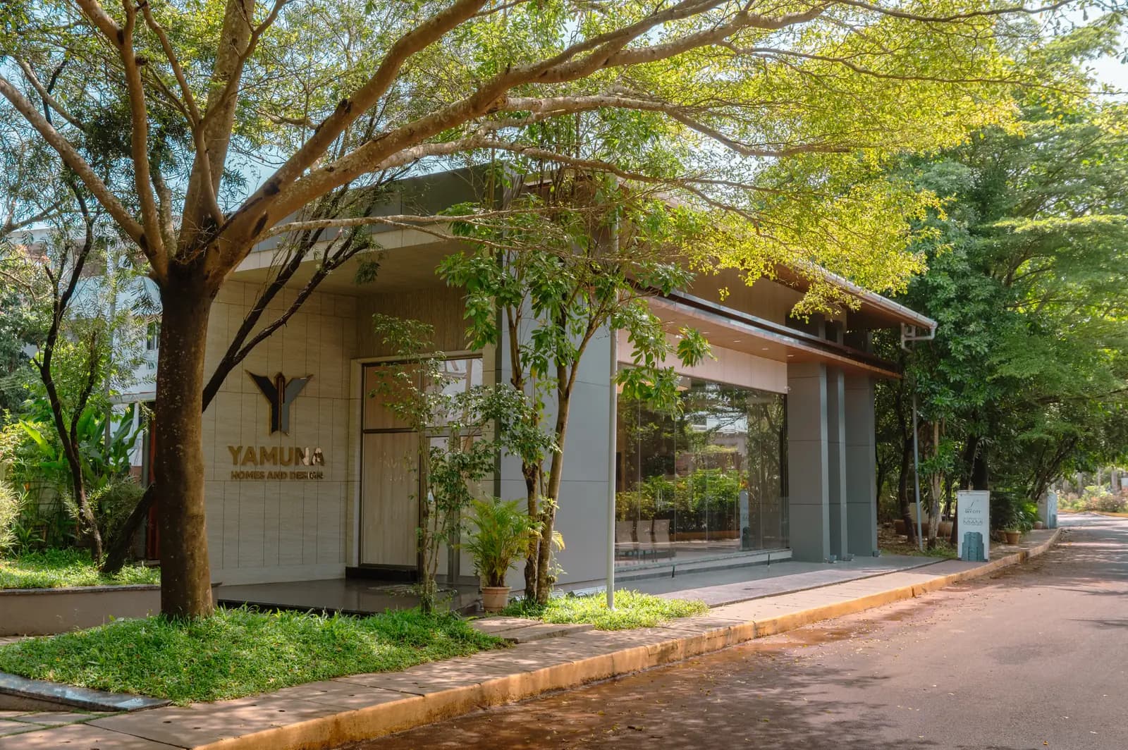A single-storey showroom on a tree-lined street, clad in pale stone under a cantilevered roof, with full-height glazing and brass lettering reading Yamuna Homes and Design